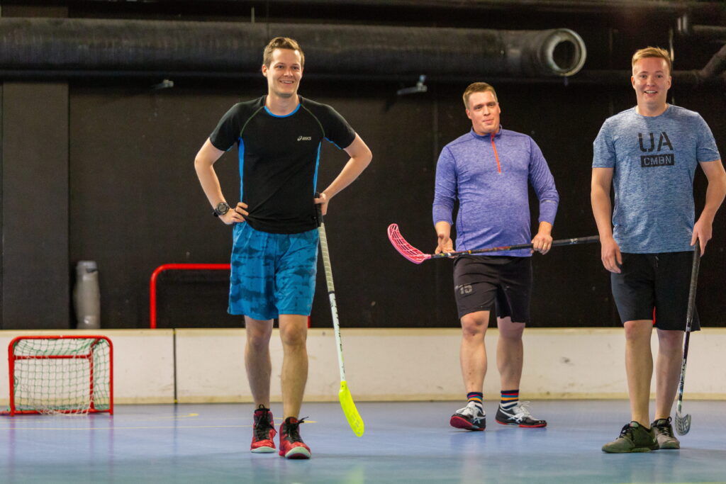 Three WeAre Solutions colleagues standing on an indoor floorball court with sticks, smiling during a team sports activity in 2018.